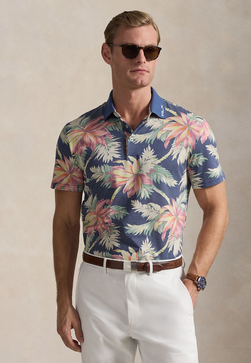 Άνδρας που φοράει σκούρα γυαλιά ηλίου, μπλε σκούρο πόλο με λουλουδάτο μοτίβο, άσπρο παντελόνι, καφέ ζώνη και μπλε ρολόι μπροστά σε ουδέτερο φόντο.