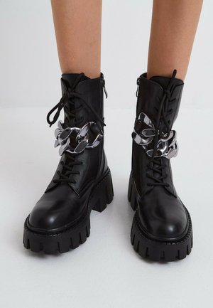 Bottes de combat noires à lacets avec semelles épaisses et volumineuses, ornées d'une chaîne argentée oversize brillante, portées sur des jambes nues sur fond blanc.