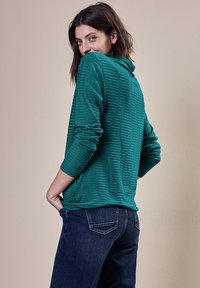 Türkise Longsleeve-Bluse mit strukturiertem Streifendesign, lockerer Passform und Rundhalsausschnitt, kombiniert mit dunkelblauen Jeans.