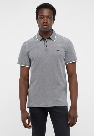Mann trägt ein graues kurzärmeliges Poloshirt mit dunklen Knöpfen und einem kleinen Logo auf der linken Brust, kombiniert mit schwarzen Hosen.