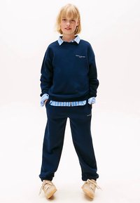 Granatowa bluza z białym napisem, nałożona na jasno-niebieską koszulę w paski. Do tego dopasowane granatowe spodnie dresowe i beżowe trampki.