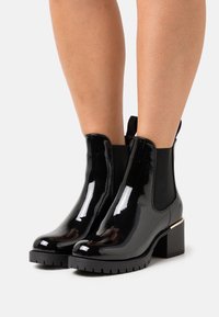 Černé lesklé kotníkové boty s elastickými bočními panely, robustní černou podrážkou a zlatým akcentem na patě. Hladká textura a moderní design.