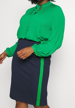 Femme portant un chemisier vert à manches longues avec des plis et une jupe bleu marine avec une bande verticale verte sur le côté.