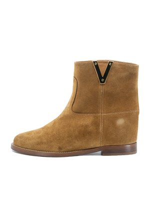 Stivale alla caviglia in suede color marrone chiaro con punta arrotondata, cuciture minime e un accento a forma di V dorato sul retro. Tacco in legno piatto.