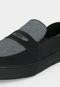 Baskets noires sans lacets avec un panneau supérieur gris texturé et une semelle en caoutchouc noire, avec un petit logo de la marque sur la bride supérieure.