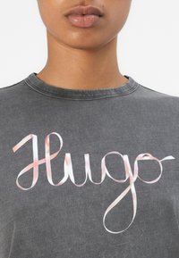 Siva pamučna majica sa rebrastim izrezom, koja sadrži veliki, stilizovani "Hugo" logo u ružičastom i belom slogu.
