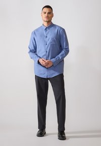 Chemise bleu à manches longues en mélange de coton avec poche avant et boutons noirs, assortie à un pantalon noir et des chaussures noires brillantes.