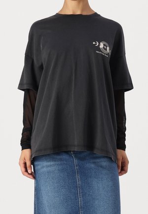 Person iført en løs sort T-shirt over en langærmet sort mesh-top og en blå denim nederdel, med en lille grafik og tekst på brystet.