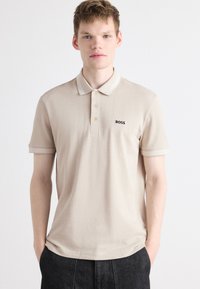 Beige Poloshirt mit klassischem Kragen, ausgestattet mit einer Dreiknopf-Leiste und dezenten Streifenakzenten an den Ärmeln. Besticktes "BOSS"-Logo.