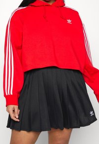 Hoodie rosso corto con design a tre strisce bianche e logo sul petto, abbinato a una gonna plissettata nera con un logo all'orlo.