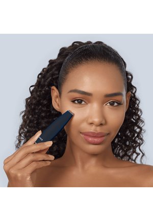 Kvinde med krøllet hår holder en mørkeblå ansigtsenhed blidt mod sin højre kind og demonstrerer hudbehandling.