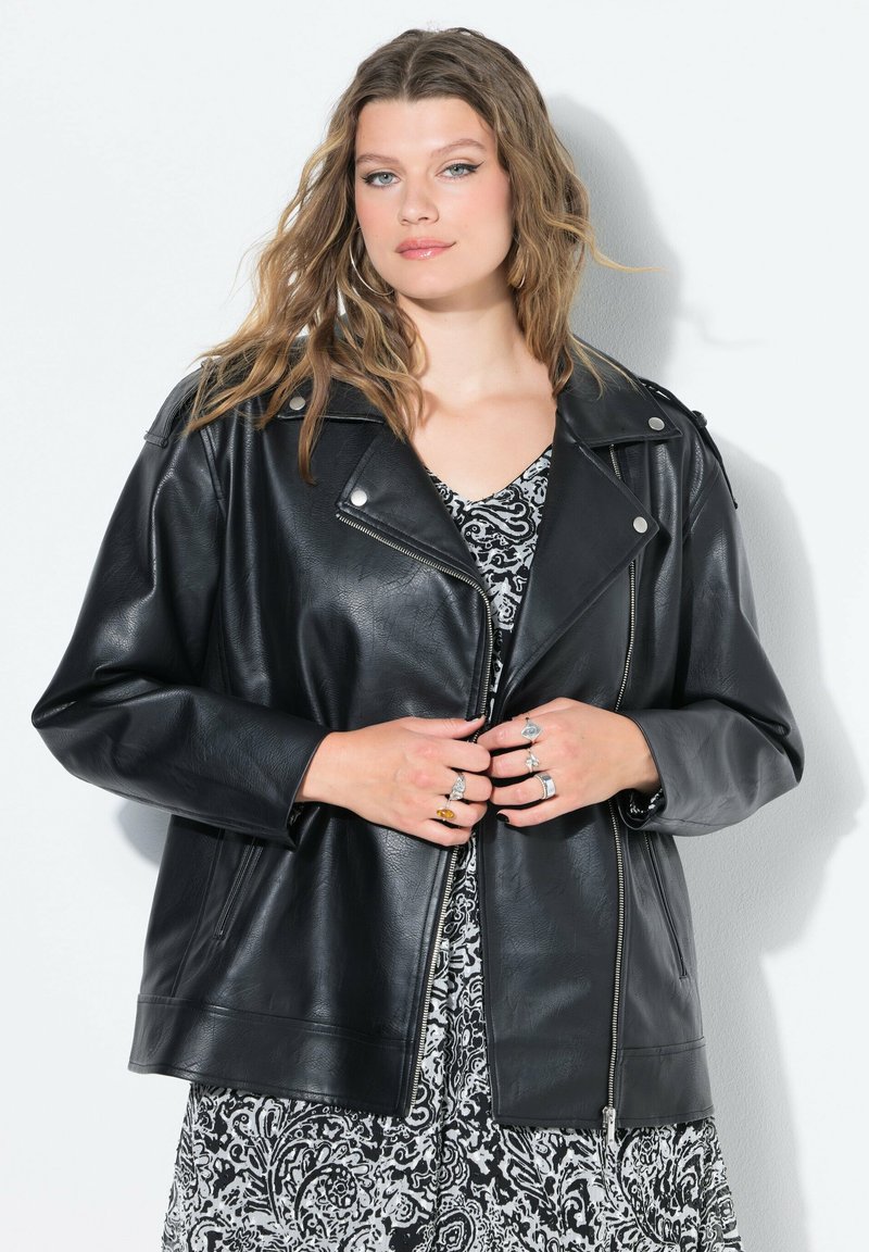 Chaqueta de cuero negra de motociclista con un corte oversize, cierre con cremallera, grandes solapas y herrajes plateados. Llevada sobre un vestido estampado.