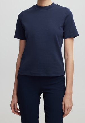 Personne portant une chemise cintrée unie bleu marine à manches courtes et un pantalon bleu marine assorti sur un fond gris clair.