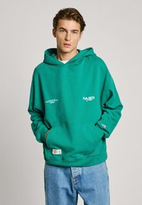 Sudadera verde con bolsillo tipo canguro, con logotipo blanco y un ajuste relajado, combinada con jeans de mezclilla de color azul claro.