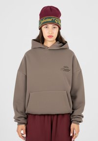 Haut de hoodie oversize marron avec une poche frontale et un logo brodé, accompagné d'un bonnet bordeaux avec une bande verte et jaune.