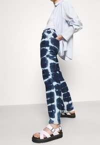 Blå tie-dye denimjeans med avslappnad passform, hög midja och sidofickor. Matchas med vita remsandaler och en ljusblå skjorta.