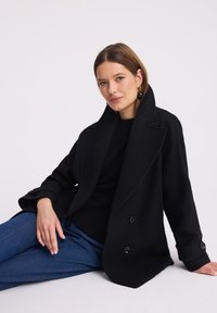 Czarny, dwurzędowy płaszcz z miękkiego materiału z szerokim kołnierzem i zapinanymi mankietami, połączony z czarnym swetrem i niebieskimi dżinsami.