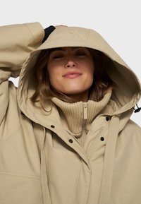 Beige hættejakke med en tyk ribbet beige krave, lynlås lukning og synlige knapper, der har et afslappet, oversized fit.