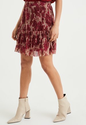 Knælængde plisseret nederdel i rødt og beige abstrakt mønster kombineret med cremefarvede ankelstøvler med høje hæle på en stående person.