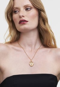Gold pendant necklace with an oval shape, featuring a decorative clasp and engraved details, worn over a black strapless top.