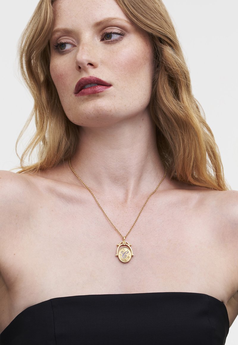Gold pendant necklace with an oval shape, featuring a decorative clasp and engraved details, worn over a black strapless top.