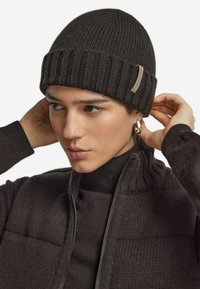 Mörk ribbad stickad mössa gjord av ull, med en vikbar kant och en liten etikett på sidan. Modellen justerar mössan i en varmtonad outfit.