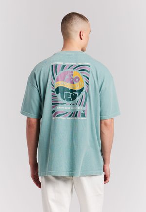 Man met de rug naar de camera, draagt een licht turquoise T-shirt met een kleurrijk abstract surflogo en de tekst "Happy Surf Since 1993", gecombineerd met witte broek.