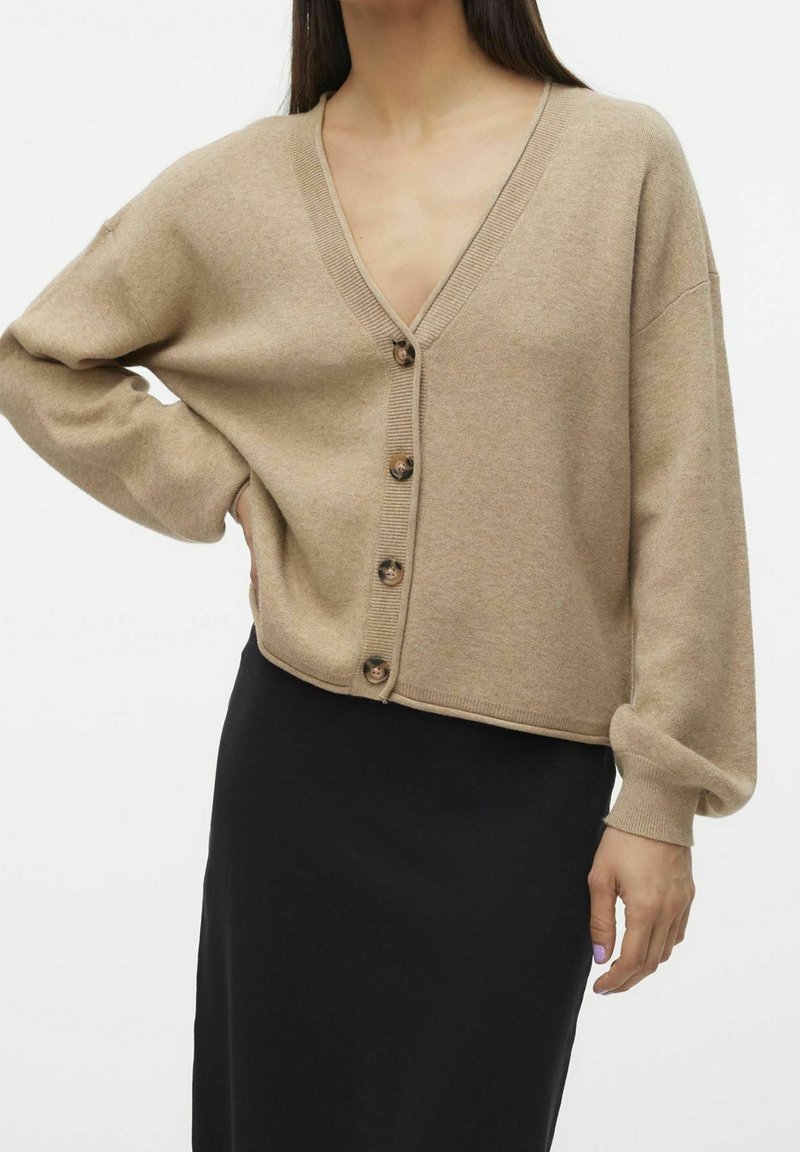 Cardigan beige en maille avec col en V, épaules tombantes et boutons en bois. Longueur courte et raccourcie avec poignets et ourlet côtelés.