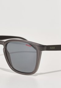 Ochelari de soare din plastic gri, cu formă pătrată, lentile întunecate și o braț texturat negru, având un logo verde "HUGO".