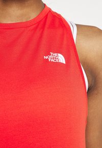 Persona con una camiseta sin mangas roja deportiva con el logotipo blanco de "The North Face" en el pecho y una correa blanca visible por debajo.
