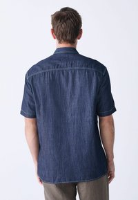 Camisa de mezclilla de manga corta en azul oscuro con cuello abotonado, dobladillo recto y costuras en contraste. Textura suave, ajuste relajado.