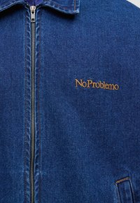 Veste en jean avec fermeture éclair argentée, col et texte brodé "No Problemo" en fil marron sur la poitrine.