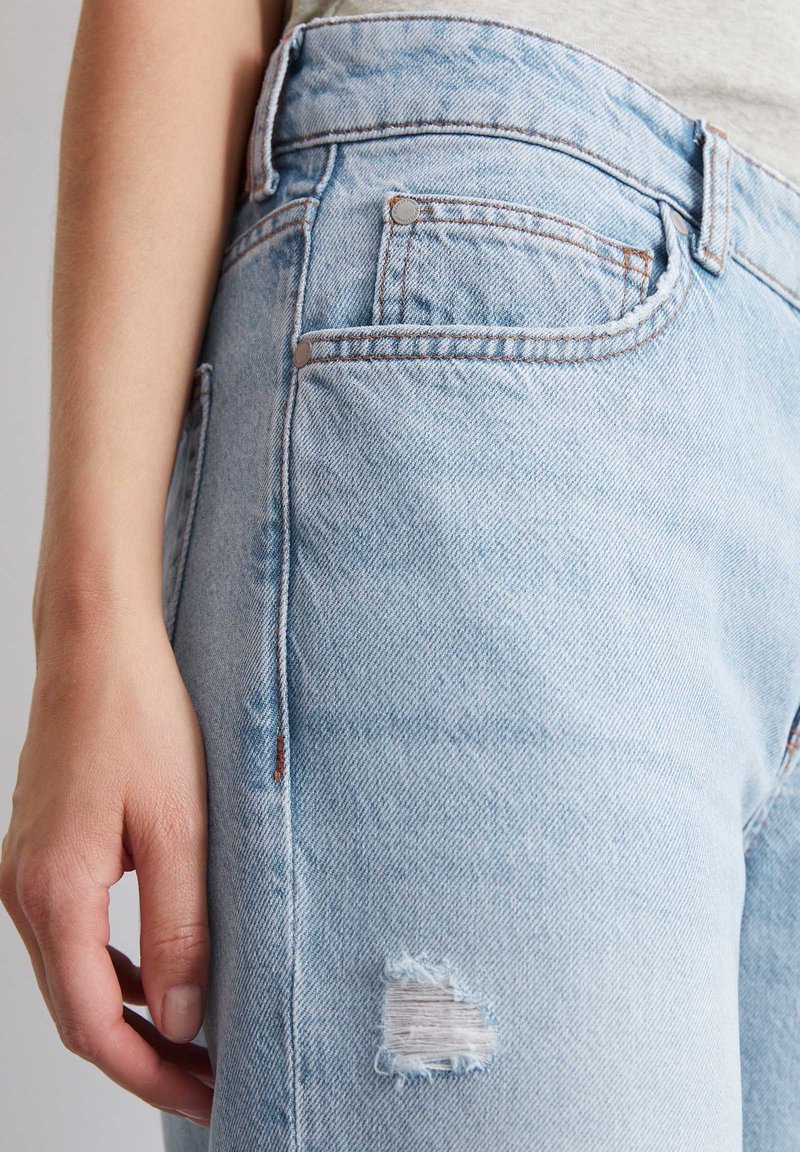 Jasnoniebieskie, znoszone dżinsy z wysokim stanem, z rozdartym kolanem, przednimi kieszeniami i kontrastowym przeszyciem wzdłuż szwów.