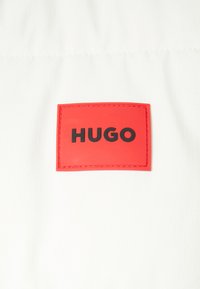 Weißer Stoff mit einem rechteckigen roten Etikett, auf dem der prägierte Text "HUGO" in fetten schwarzen Buchstaben steht. Glatte Textur mit einem subtilen Gittermuster.