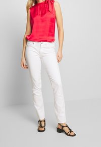 Haut sans manches en satin rouge avec un nœud décoratif à l'épaule, associé à un jean blanc ajusté et des sandales noires à lani ères. Texture lisse.