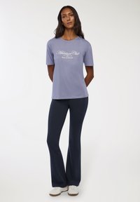 T-shirt corto a maniche corte color lavanda chiaro con la scritta "Athleisure Club Relax & Wellness" stampata in bianco. Abbinato a pantaloni blu navy a sigaretta e scarpe da ginnastica bianche.