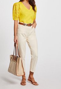Gul v-ringad topp med spetspuffärmar, beigefärgade högmidjade byxor, brun bälte, bruna sandaler och en ljus tote-väska med läderdetaljer.
