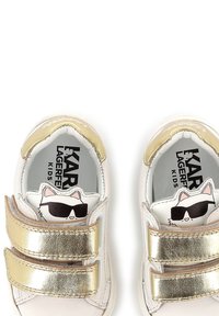 Baskets blanches pour enfants avec des bandes Velcro métallisées dorées, ornées d'un graphique de chat sur la semelle intérieure. Texture en cuir lisse, design à bout rond.