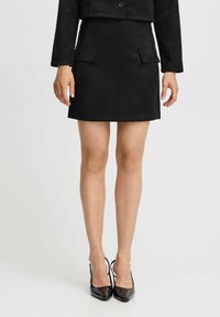 Schwarzer Wildleder-Minirock mit gerader Silhouette, ausgestattet mit zwei flachen frontalen Taschen und einer glatten Textur. Kombiniert mit schwarzen Lackleder-Pumps.