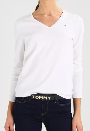 Femme portant un haut blanc à manches longues et col en V ainsi qu'un jean foncé avec une ceinture noire étiquetée "TOMMY" à la taille.
