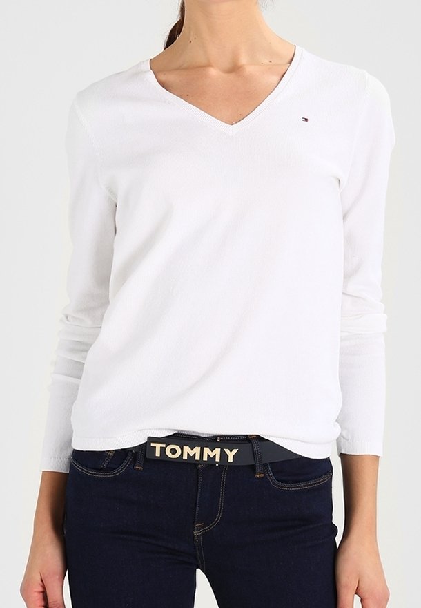 Femme portant un haut blanc à manches longues et col en V ainsi qu'un jean foncé avec une ceinture noire étiquetée "TOMMY" à la taille.