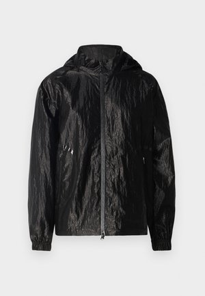 Zwarte, gestructureerde, lichte jas met een capuchon en ritsluiting. Voorzien van elastische mouwboorden en twee zijzakken met ritssluiting.