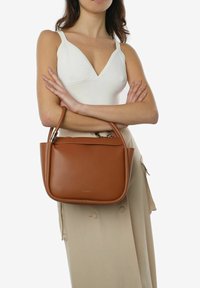 Brązowa skórzana torebka o uporządkowanym kształcie, z dwoma krótkimi uchwytami i minimalistycznym designem. Teksturowane wykończenie, bez widocznych logo.