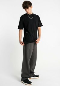 Czarny bawełniany t-shirt z okrągłym dekoltem i krótkimi rękawami; połączony z luźnymi szarymi spodniami; model stoi z dłońmi w kieszeniach.