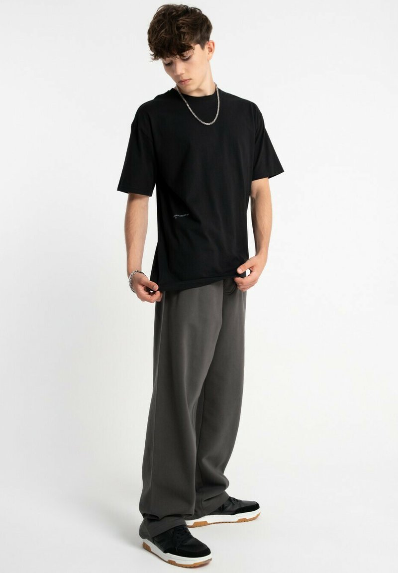 Czarny bawełniany t-shirt z okrągłym dekoltem i krótkimi rękawami; połączony z luźnymi szarymi spodniami; model stoi z dłońmi w kieszeniach.