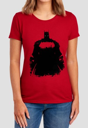 Woman wearing red T-shirt with black Batman silhouette and city skyline on the chest, paired with blue jeans.