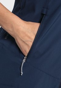 Námořnická kapsa na zip s šedou táhlem na hladké, texturované tkanině. Částečně je vidět ruka, jak sahá do kapsy.