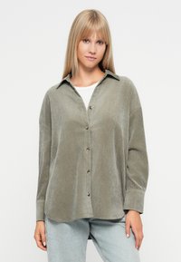 Zielona koszula z korduroju z guzikiem z przodu, kołnierzykiem i długimi rękawami, charakteryzująca się luźnym krojem i gładką fakturą. Noszona na białej koszuli.