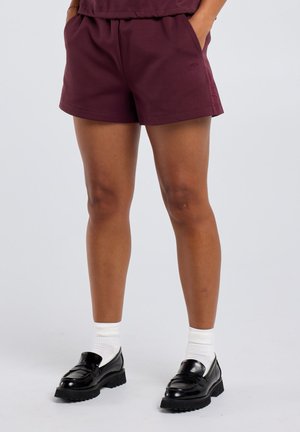 Persoon draagt bordeauxrode korte broek, witte crew-sokken en zwarte leren loafers, staand met handen in de zakken tegen een lichte achtergrond.