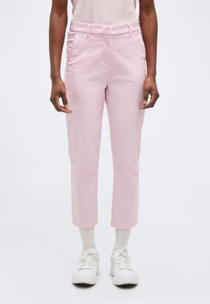 Persoon draagt lichtroze op maat gemaakte broek, witte geribbelde sokken en witte veterschoenen, staand tegen een effen achtergrond.